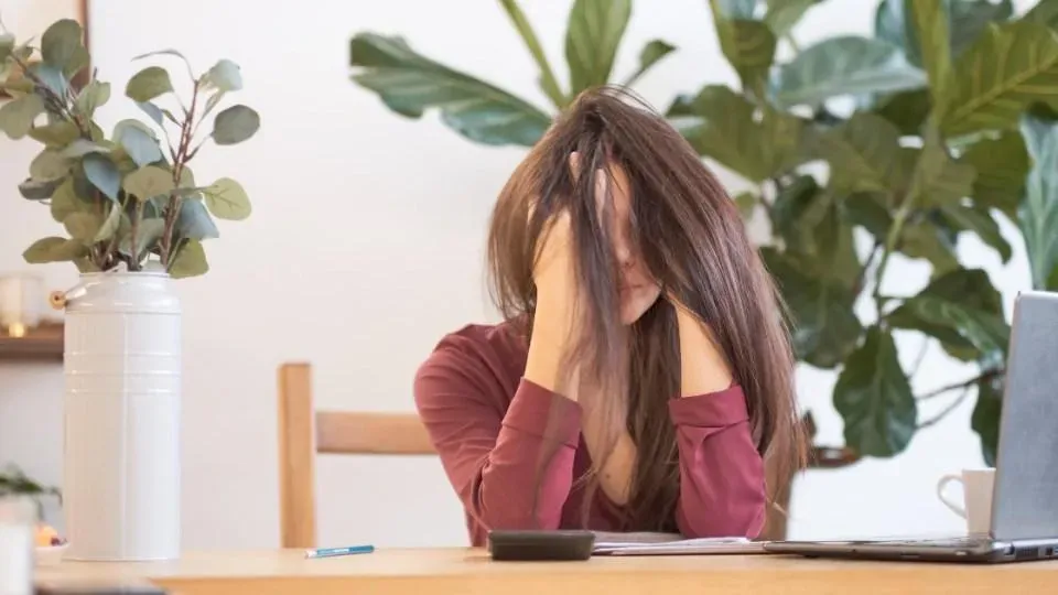 Woman sitting at a desk with her hands over her face looking really stressed. Her hair is a mess and she has work in front of her., Apa Itu Hustle Culture?, Ciri-ciri Hustle Culture, Dampak Buruk Hustle Culture, Tips Mengatasi Hustle Culture