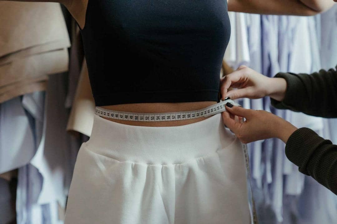 1. Sebelum mengambil ukuran, pastikan kamu memakai baju yang tipis atau cropped top. Untuk tahu ukuran lingkar pinggang dengan mudah, gunakan pita meteran., 2. Kalau nggak punya pita meteran, tali juga bisa jadi andalan. Lilitkan tali di sekitar pinggang dan potong seukuran dengan lingkar pinggang. Kemudian ukur panjang tali yang sudah dipotong menggunakan penggaris., 3. Nggak ada tali, pakai ikat pinggang juga bisa kok. Lakukan cara yang sama dan usahakan untuk rileks saat mengambil ukuran, jangan menggembungkan perut atau menarik ke dalam., 4. Manfaatkan peralatan yang ada di rumah. Ambil celana yang pas dengan ukuranmu, kemudian letakkan pada permukaan datar. Kemudian ukur bagian lingkar pinggang pada celana tersebut dengan penggaris., 5. Cukup dengan tangan kosong juga bisa, lho. Ambil ukuran dengan cara menghitung total jengkal jari pada lingkar pinggangmu. Satu jengkal jari biasanya setara dengan 22,86 cm.