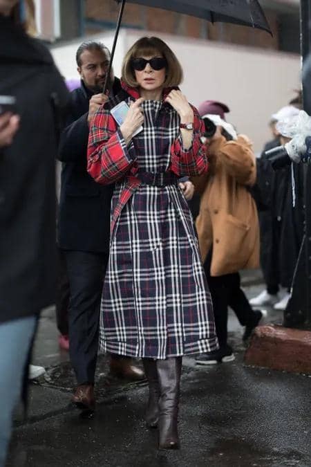 4f10e4962429db04d8b8125ca78310dfb0-anna-wintour-13.rdeep-vertical.w450.jpg, 1. Rambut bob dan kacamata hitam udah jadi ciri khas Anna sejak dulu. Seperti saat hadir di CFDA Awards Dinner tahun 1999 dengan gaun satin pink-nya., 2. Selalu jadi sosok penting di balik Met Gala, ini salah satu penampilan Anna di Met Gala 2017 memakai gaun Chanel dengan detail rok feathery., 3. Menemani Ratu Elizabeth II saat menonton fashion show, Anna tetap konsisten dengan kacamata hitamnya. Ia pakai dress floral dengan boots yang chic., 4. Anna tahu cara bermain dengan warna dan motif. Kali ini ia memadukan dress dan outer plaid dengan boots yang bikin gayanya stylish dan edgy., 5. Di Met Gala 2024, Anna datang pakai tuxedo coat rancangan Loewe dengan hiasan bunga tulip yang terinspirasi dari Charles Frederick Worth 1889