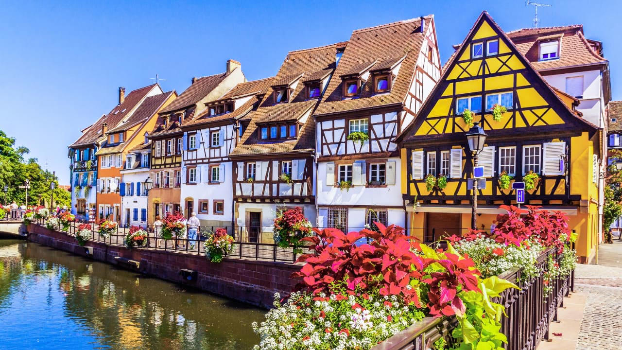 3. Colmar, France Colmar does not ask for attention—it commands it. With its riot of half-timbered homes, canal reflections, and Alsatian flourishes, it walks the line between theatre and truth. Even its shadows are deliberate. (Image: Canva) 1. Hallstatt, Austria Balanced between lake and limestone, Hallstatt reads like a forgotten chapter from an alpine fable. Its salt-stained history, mirrored waters, and slate-roofed homes draw quiet reverence, not crowds. In winter, it feels paused; in summer, painted. (Image: Canva), 2. Shirakawa-go, Japan Cradled by mountains and wrapped in thatch, Shirakawa-go holds firm to tradition. Its steep-roofed farmhouses, designed to endure snow and time, glow amber at dusk. The seasons arrive like guests—welcomed, noticed, remembered. (Image: Canva), 3. Colmar, France Colmar does not ask for attention—it commands it. With its riot of half-timbered homes, canal reflections, and Alsatian flourishes, it walks the line between theatre and truth. Even its shadows are deliberate. (Image: Canva), 4. Bibury, England Set beside the Coln and stitched into the hills, Bibury whispers of England before speed. Honey-coloured cottages in tight rows, trout streams that glisten without reason, and hedgerows that seem older than maps. (Image: Canva), 5. Giethoorn, Netherlands There are no cars in Giethoorn. Only boats, bridges, and hushed conversations. It is a village that floats, slowly, in its own rhythm. The canals speak softly; the thatched roofs never hurry. (Image: Canva), 6. Reine, Norway Beyond the Arctic Circle lies Reine—a scattering of crimson cabins that appear borrowed from a painting. Mountains rise like frozen waves behind them, and the sea never stops watching. Reine is remote, but never empty. (Image: Canva), 7. Eze, France High above the Riviera, where the cliffs fall into blue, sits Eze—a village of stone and shadow. Its alleys twist upward as if climbing toward memory. Time lingers here, in the scent of thyme and sea. (Image: Canva), 8. Goreme, Turkey Caves carved by wind and wonder—Goreme belongs to the Earth, not to man. Fairy chimneys pierce the sky, and hot-air balloons rise like thoughts at dawn. It is a village of both ground and sky. (Image: Canva), 9. Dinant, Belgium Hemmed in by cliff and river, Dinant feels composed rather than built. Saxophones hang from bridges, limestone watches from above, and the Meuse flows through it like a quiet refrain. Gothic never looked so lyrical. (Image: Canva), 10. Leavenworth, Washington, USA This village is not a replica—it’s a reinvention. Bavarian beams beneath American skies, brats beside apple pie. Surrounded by pine, performance, and peaks, Leavenworth is both celebration and curiosity, wrapped in wood and wonder. (Image: Canva)