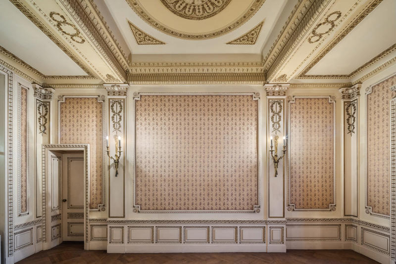 The libraries, reception rooms, and parlors have retained their Gilded Age opulence with decorated walls and ceilings. The five-story townhouse is located across from the Museum of Modern Art., The entry gallery on the first floor features wood paneling throughout., Neo-Georgian architectural touches include columns and decorative molding known as cornices., Two staircases lead to the upper floors., Rooms on the upper floors feature floor-to-ceiling windows and Juliet balconies., The dining room is shaped like an octagon.