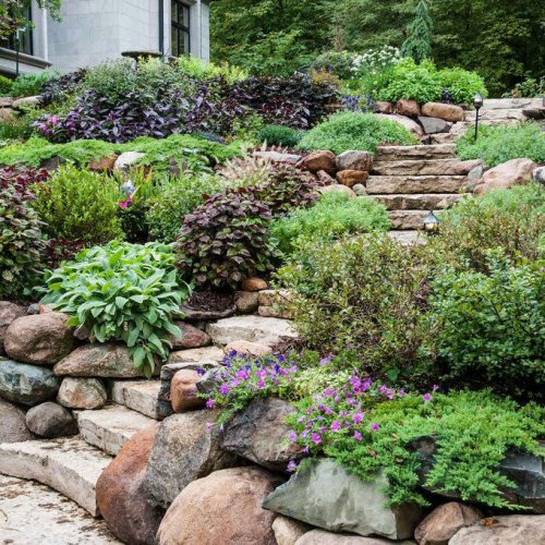 Lush, multicolored plants and flowers surround stone steps rising through a landscaped garden, creating a serene outdoor pathway., Terraced Hillside Landscaping, Stone steps lead up through a landscaped garden, surrounded by greenery and decorative plants, towards a wooden fence and small shed in the background., Steps with Retaining Wall, A two-story house with large windows features a deck and is surrounded by lush greenery and rocks under a clear blue sky., Create a Rock Garden, Adirondack chairs surround a fire pit, facing a tiered stone waterfall, set in a landscaped backyard near a green house., Hillside Water Feature, A large, red-brick house features intricate stonework and multiple gabled roofs, surrounded by well-maintained landscaping and a clear blue sky., Front Yard Retaining Wall, Six colorful chairs surround a circular fire pit. A dock and water are visible in the background, surrounded by lush green grass and bushes., Patio Overlook, A patio features wicker furniture arranged around a decorative rug, with surrounding landscaped gardens, trees, and a clear blue sky above., Hillside Landscaping with Mulch, A large dining table with black wicker chairs sits under a beige umbrella, surrounded by lush greenery and colorful plants in a sunny outdoor setting., Tiered Raised Beds, A modern house with a multi-pitched roof and colorful facade stands beside a driveway, surrounded by greenery and a clear blue sky., Front Deck for a Sloped Yard, Lush green lawn features a stone retaining wall adorned with colorful flowers in pots, surrounded by tall trees under clear blue skies., Prairie Garden Hillside Landscaping, FAQ, Top 10+ Hillside Landscape Ideas to Make the Most of Your Sloped Yard